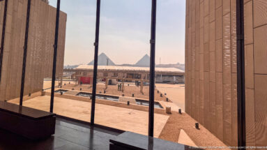 Giza pyramids seen through tall glazing from inside the Grand Egyptian Museum, beyond the museum forecourt.