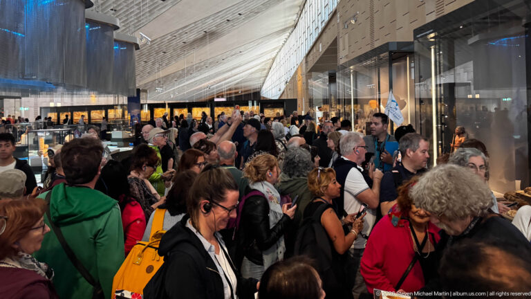 Crowded Grand Egyptian Museum gallery with visitors, glazed walls, display cases, and a high angular ceiling.