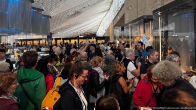 Crowded Grand Egyptian Museum gallery with visitors, glazed walls, display cases, and a high angular ceiling.
