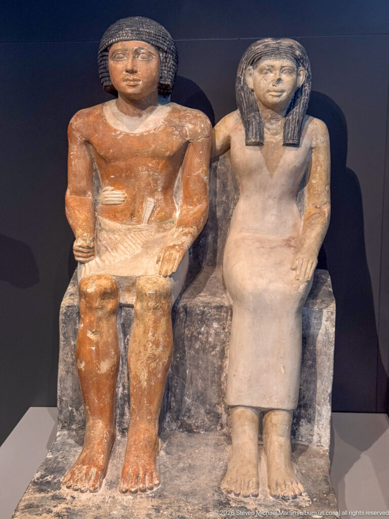 Seated painted stone statues of a man and woman shown frontally on a shared block-like seat.
