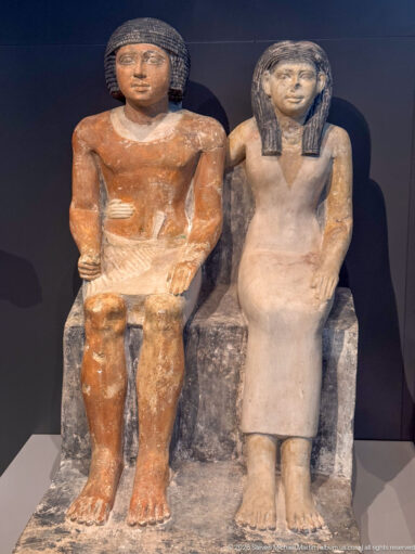 Seated painted stone statues of a man and woman shown frontally on a shared block-like seat.