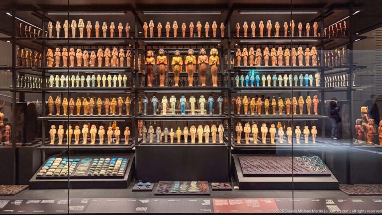 Large museum wall display packed with rows of shabtis and funerary figures, with smaller amulet trays in the foreground.