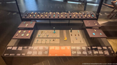 Overhead museum display of many ancient Egyptian amulets grouped by type on dark panels, including ankhs and small figures.