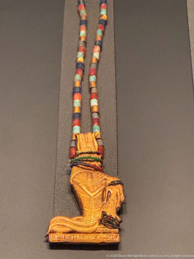 Multicolored cylindrical bead strand with a gold falcon-shaped pendant showing a hooked beak and folded wing.