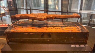 Dark rectangular funerary container with orange-brown surface patches beneath a long weathered wooden element mounted horizontally.