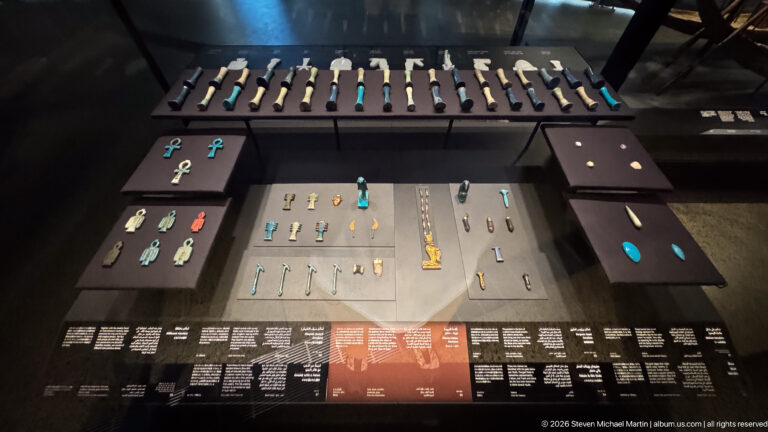 Display case with many small Egyptian amulets and pendants arranged on dark panels, including ankhs, figures, and turquoise pieces.