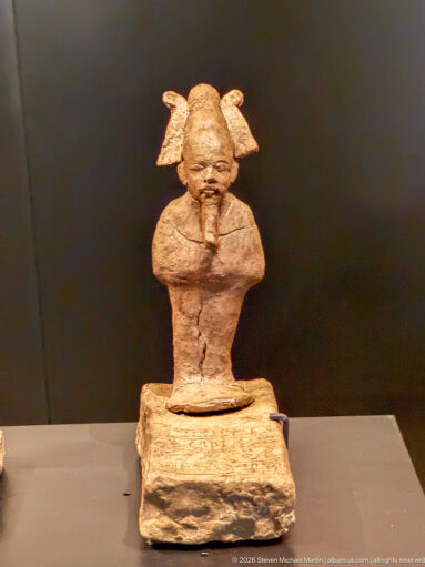 Small weathered seated or kneeling figure with broad projecting ears, compact body, and strongly modeled face on a block base.