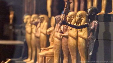 Oblique side view of a row of small Egyptian deity figures, with a dark foreground figure extending one arm.