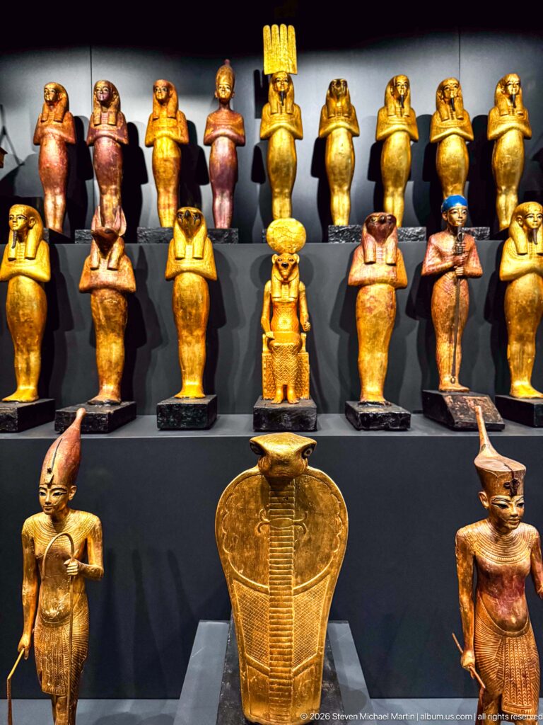 Tiered display of many gilded and painted Egyptian deity figures, including human- and animal-headed forms around a large central emblem.