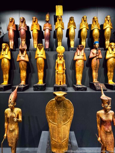 Tiered display of many gilded and painted Egyptian deity figures, including human- and animal-headed forms around a large central emblem.