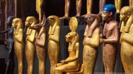 Close grouping of gilded Egyptian deities with falcon, canine, and human heads, including one seated figure beneath a solar disk.