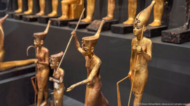 Several gilded standing deity figures with tall crowns, staffs, and different poses arranged closely in a museum case.