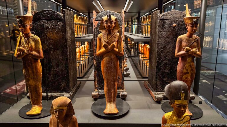 Standing gilded and painted funerary figures displayed before tall dark coffins in a densely installed museum gallery.