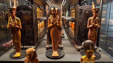 Standing gilded and painted funerary figures displayed before tall dark coffins in a densely installed museum gallery.
