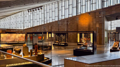 Wide view along the Grand Egyptian Museum’s main gallery, with glass vitrines and tall stone walls forming a bright, open circulation space.
