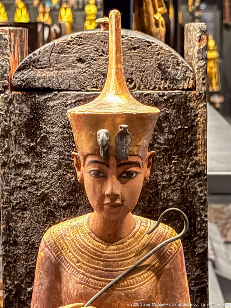 Close-up of gilded anthropoid figure with tall headdress, broad collar, dark outlined eyes, and a curved object crossing the torso.
