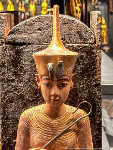 Close-up of gilded anthropoid figure with tall headdress, broad collar, dark outlined eyes, and a curved object crossing the torso.
