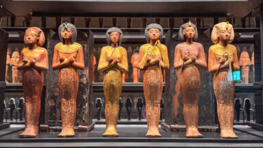 Tightly packed rows of small blue-green and neutral-toned mummiform shabti figures displayed in a museum case.