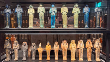 Two-tier museum display filled with many shabti figures in turquoise, cream, ochre, black, and painted finishes.