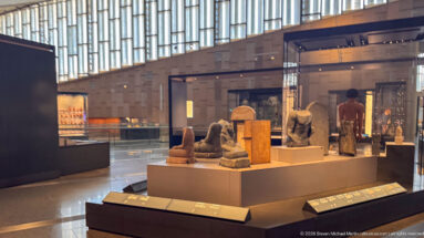 Glass case holding multiple small gilded wood ancient Egyptian statuettes on stepped platforms in the Grand Egyptian Museum gallery.