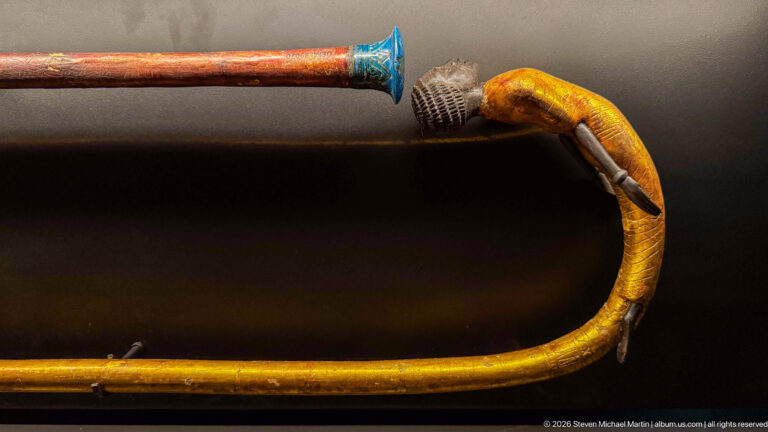 Yellow-gold curved cane ending in a bird head with projecting beak and dark feather details, beside a staff with blue fitting.