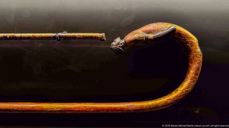 Long curved ancient cane with painted animal-head terminal, pointed ears, elongated snout, and reddish-gold decorated shaft.