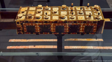 Decorated rectangular game board with a square grid, many spool- and cone-shaped playing pieces, and patterned side panels.