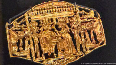 Embossed gold plaque with a large seated figure beneath a canopy, flanked by smaller standing attendants and decorative elements.