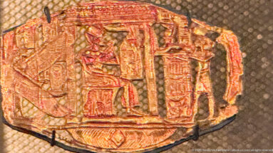 Small embossed gold plaque with multiple standing figures beneath a shrine-like architectural frame and traces of red color.