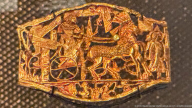Small embossed gold plaque with chariot wheel, human figures, and overlapping animals in a dense narrative scene.