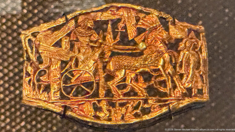 Small embossed gold plaque with chariot wheel, human figures, and overlapping animals in a dense narrative scene.