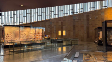Curved circular metal and glass fixture suspended in a Grand Egyptian Museum gallery, viewed from below against softly lit walls.