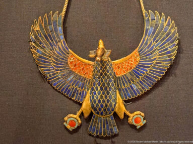 Gold and blue inlaid bird with fully spread wings, red shoulder bands, scale-patterned body, and small hanging circular ornaments.