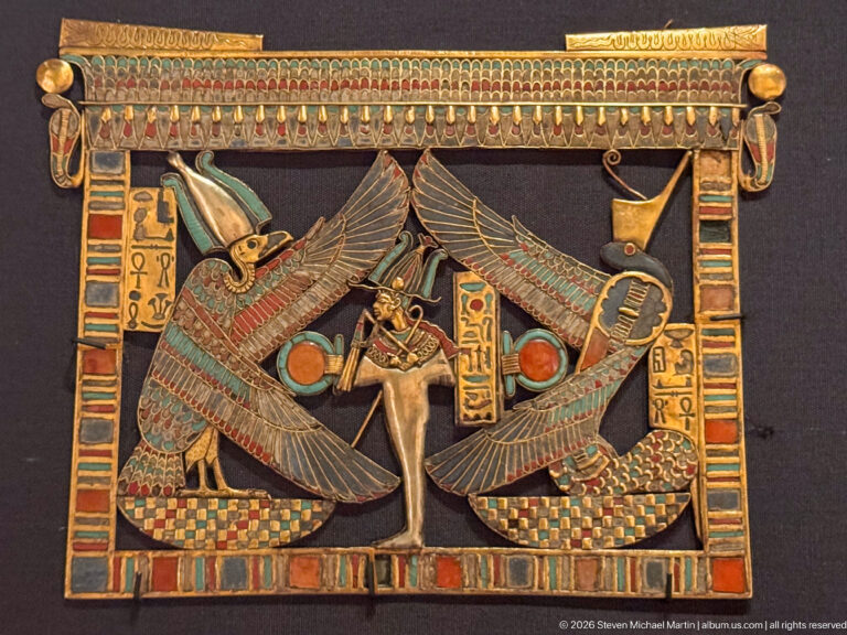Rectangular gold pectoral with central standing figure, two opposing winged deities, hieroglyphs, solar disks, and multicolored inlay.