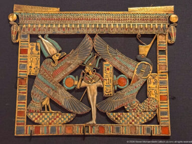 Rectangular gold pectoral with central standing figure, two opposing winged deities, hieroglyphs, solar disks, and multicolored inlay.
