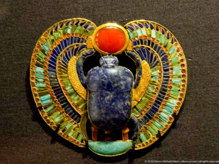 Circular inlaid ornament with dark blue scarab, red solar disk, and sweeping turquoise-and-gold wings forming a ring.