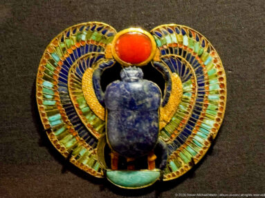 Circular inlaid ornament with dark blue scarab, red solar disk, and sweeping turquoise-and-gold wings forming a ring.