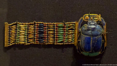 Broad multicolored beaded bracelet ending in a large blue scarab set within an ornate gold frame.