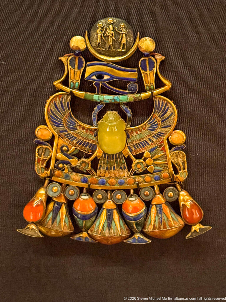 Ornate gold pectoral with solar disk, winged scarab, Eye of Horus motif, and multicolored inlaid pendant forms.