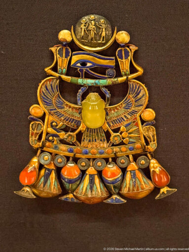 Ornate gold pectoral with solar disk, winged scarab, Eye of Horus motif, and multicolored inlaid pendant forms.