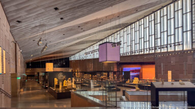 Wide Grand Egyptian Museum exhibition hall with high ribbed ceiling, long clerestory windows, suspended screens, and rows of display cases.
