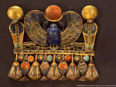 Gold inlaid pectoral with blue winged scarab, red solar disk, flanking cobras, and rows of multicolored pendant elements.