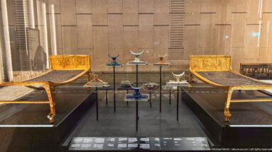 Pair of gilded beds or couches facing inward across a museum display with slender stands and small ceremonial objects between them.