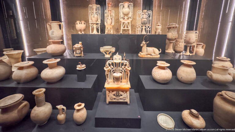 Tiered museum display containing many stone and ceramic vessels, small figures, and ornate polychrome luxury objects.