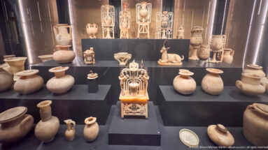 Tiered museum display containing many stone and ceramic vessels, small figures, and ornate polychrome luxury objects.