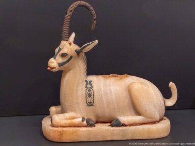 Polychrome recumbent ibex or gazelle with long curved horns, dark facial markings, curled tail, and hieroglyphic emblem on its flank.