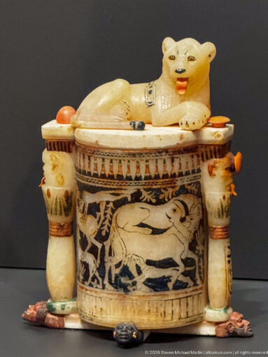 Recumbent cream-colored lion atop a decorated cylindrical vessel flanked by two smaller containers with painted ornament.