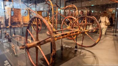 Ancient wooden two-wheeled chariot with large spoked wheels, long pole, curved frame, and surviving painted or patterned details.