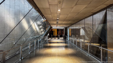 Long Grand Egyptian Museum corridor with glass railings, angular stone walls, ceiling lights, and a dark doorway at the far end.