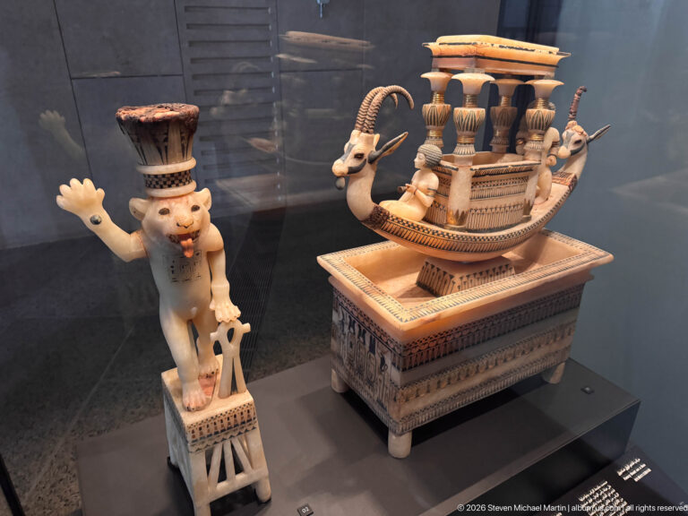 Display with an ornate boat-shaped polychrome object beside a standing dwarf-like figure wearing a tall headdress and raising one arm.